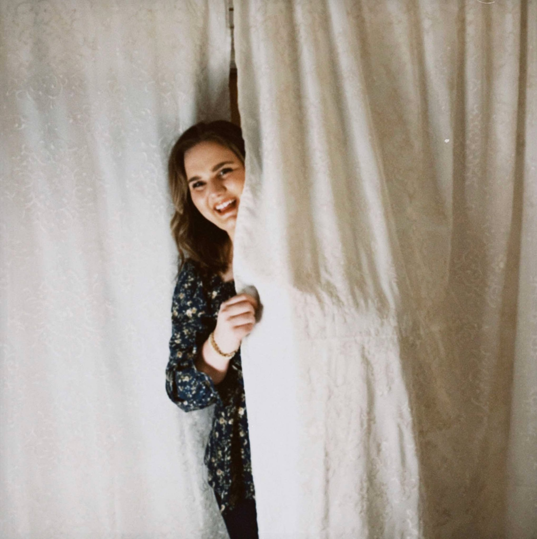 Shayna Hansen peeking playfully from behind white curtains, representing curiosity and storytelling in web design for simple living brands.